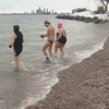 Trois personnes en costumes de bain, les pieds dans l'eau alors que la neige recouvre  une rive proche.