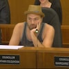 Un homme est assis dans un fauteuil devant un micro au cours d'une réunion. Il porte un chapeau et un débardeur.