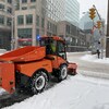 Un petit camion orange s'affaire à déneiger les trottoirs d'un coin de rue enneigé à Ottawa. 
