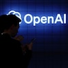 Devant le logo d'OpenAI, des personnes regardent leur téléphone.