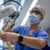 Une chirurgienne portant des vêtements de salle d'opération fait une démonstration en utilisant un bras robotique équipé d'un genre de perceuse. Elle approche l'outil d'une réplique d'une articulation de la jambe.