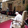Le pape assis seul à une chaise, à plusieurs mètres de plusieurs personnes habillées en vêtements traditionnels.