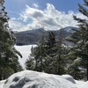 Derrière des sapins, on voit un lac gelé au pied d'une montagne sillonnée par des pistes de ski.