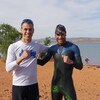 Deux athlètes posent devant un bassin artificiel au coeur du désert de St. George, en Utah. Ils sourient, chacun ayant un poing fermé en signe de victoire. L'un d'entre eux, le professionnel, porte une combinaison et un casque de natation. L'autre homme à sa gauche, Patrick Dumont, est en chandail et short.