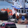 Des travailleurs sur le quai manipulent des équipements de pêche. En arrière-plan : un bateau.