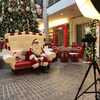 Le père Noël est assis sur un petit sofa de deux places, entouré d'un décor de Noël. Devant lui est installé une appareil photo.