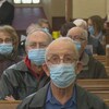 Des gens sont photographiés de face alors qu'ils sont assis à l'église avec des masques bleus au visage. 