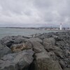 Gros-plan sur un enrochement en bordure du fleuve. Au loin, on voit le phare de Pointe-au-Père et quelques maisons résidentielles.