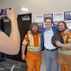 Un homme en habit est entouré de deux hommes vêtus d'un uniforme de travail fluorescent. Ils sourient en se faisant prendre en photo.