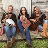 Trois femmes assises devant une grange avec leur instrument de musique à la main. 