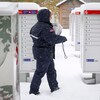 Une femme en vêtements d'hiver aux couleurs de Postes Canada marche dans la neige et s'approche de boîtes à lettres communautaires.
