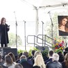 Une femme sur scène parle dans un micro, devant une foule assise lors d'un office funèbre. 