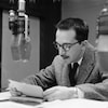 Raymond Charette lisant ses notes dans un studio de radio, derrière le micro.