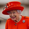 La reine porte un chapeau orange et un collier de perles et sourit. 