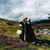 La reine Élisabeth II pose devant des collines verdoyantes, tenant un bouquet blanc dans sa main et regardant au loin. 