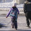Une enfant vue de dos, portant ses habits d'hiver et un sac orné d'un chat, se dirige vers l'école en compagnie d'un adulte.