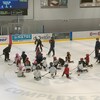 Des enfants en tenue d'hockeyeurs sont assis au centre d'une patinoire et regardent un entraîneur en train de leur montrer un mouvement avec son bâton.
