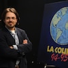 Un jeune homme se tient à côté d'une pancarte du concours fictif « La course 94-95 », montrant un globe terrestre.
