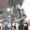 Le robot humanoïde gris se tient debout dans une salle de congrès et fait un salut avec la main levée en souriant.
