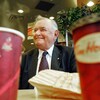 Assis à une table dans un Tim Hortons entre deux cafés et un sac de papier qui contient un beigne, Ron Joyce sourit en regardant vers sa droite.
