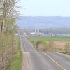 Une route droite asphaltée entourée de terres agricoles et une ferme avec des silos à grains au loin.
