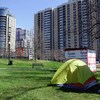 Un abri en bois et une tente dans un parc municipal, devant de hauts immeubles d'appartements.  
