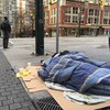 Un homme est enveloppé dans un sac de couchage posé sur des morceaux de carton sur le trottoir et tente de dormir, alors que derrière lui un piéton attend de traverser une intersection.