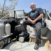 Un homme assis sur un petit bateau remorqué par une voiture. Il a un pied sur une bombonne d'air couchée sur le fond du bateau.