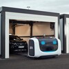 Une photo montrant le robot Stan en train de déposer une voiture dans un garage. Le robot est composé d'une mince plate-forme métallique et d'une grande boîte à l'avant contenant l'équipement électronique.