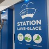 Une pompe, bleue, avec des pictogrammes et les inscriptions : « Station lave-glace », « payez », « ouvrez » et « remplissez ».