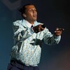 Stromae pointe le public sur scène. Il porte une chemise bleue.