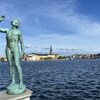 Une statue du parterre de l'hôtel de ville de Stockholm et au loin, derrière un plan d'eau, on aperçoit une partie de la vieille ville.