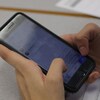 Un étudiant tient entre ses mains son téléphone cellulaire en salle de classe. Sur son bureau se trouvent des exercices scolaires.