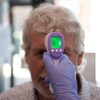 Une main couverte d'un gant de latex bleu tient un thermomètre infrarouge devant le front d'un homme aux cheveux et à la barbe blanche. Le lecteur numérique de l'appareil indique que la température corporelle de l'homme est 36,0 degrés Celsius.