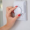 Plan rapproché de la main d'une femme aux ongles vernis rouges qui tourne la molette d'un thermostat dans un appartement.