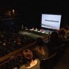 Un public regarde des personnes attablées sur la scène d'un théâtre.