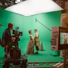 Une équipe tourne une scène de film dans un studio devant un écran vert; un acteur et une actrice habillés en costumes d'époque sont au centre. 