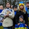 Des gens en rang manifestent dehors, enveloppés dans drapeaux bleu et jaune de l'Ukraine. Une personne exhibe une photo d'une enfant couchée dans un lit d'hôpital. 