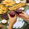 Deux personnes, dont on ne voit que les mains, trinquent avec leur verre de vin rouge. À l'arrière-plan, une table est remplie de nourriture. 