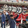 Un homme portant un complet et une casquette de champions soulevant un trophée au bout de ses bras devant des joueurs de hockey.