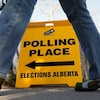 Une pancarte jaune indiquant la direction d'un bureau de vote est posée sur un trottoir. On aperçoit les jambes d'un passant qui se dirige vers ce lieu.