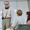 Un homme portant un casque d'écoute autour du cou discute avec une femme et un homme assis face à lui sur un banc public, pendant le tournage d'un film à Paris. 