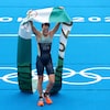 Une femme qui vient de compléter une épreuve de triathlon porte une banderolle avec le logo olympique haut dans les airs et sourit. 