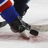Deux bâtons de hockey se disputent la rondelle entre des patins lors d'un match.