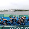 Des cyclistes passent devant le logo des Jeux de Tokyo 2020. On voit en arrière-plan la baie et un pont qui la surplombe.