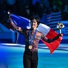 Un patineur sourit sur la glace avec une médaille de bronze au cou. Il tient un drapeau aux couleurs de la France.