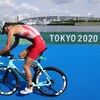 Un triathlète passe en vélo devant le logo des Jeux de Tokyo 2020 pendant l'épreuve du relais mixte. On voit un immense pont à l'arrière. 