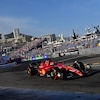 Une F1 Ferrari roule sur un circuit de F1. En arrière-plan, on voit des gradins remplis, des immeubles et des montagnes. 