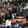 Un homme allume un grand rond d'aluminium avec la flamme olympique sur une scène, et des centaines de personnes le regardent.