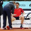 Un joueur de tennis assis en bordure du terrain grimace pendant qu'un  soigneur lui parle.
